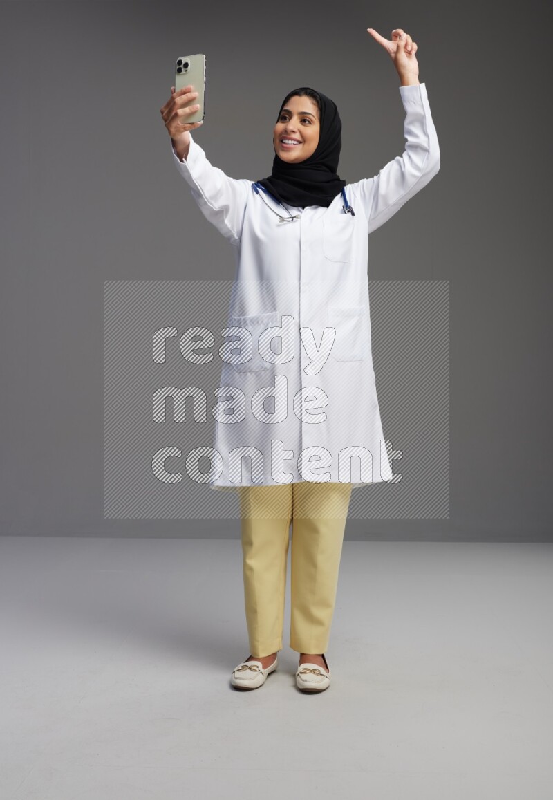 Saudi woman wearing lab coat with stethoscope standing taking selfie on Gray background