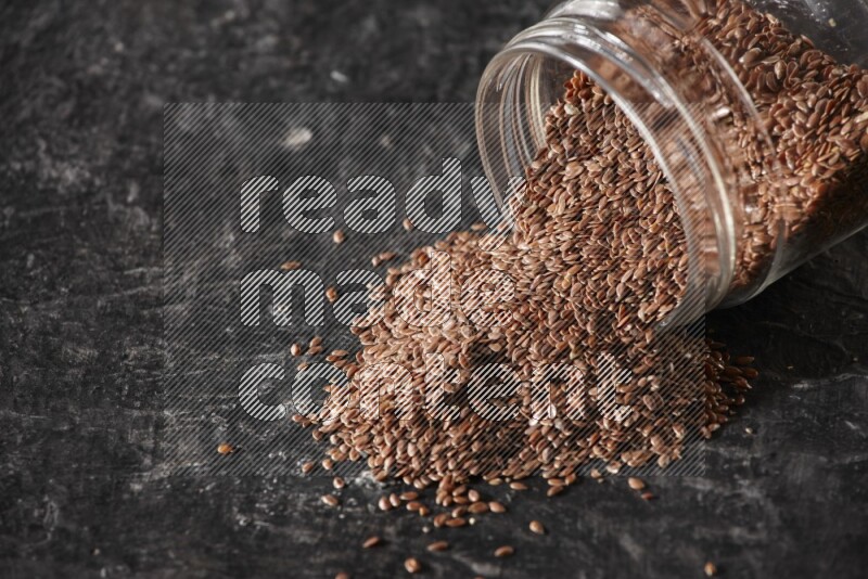 A glass jar full of flaxseeds flipped and seeds spread out on a textured black flooring