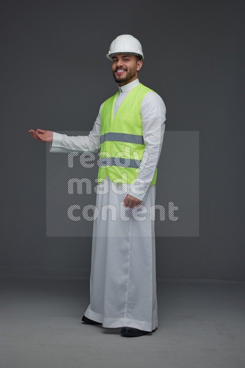 A Saudi man wearing Thobe with a yellow safety vest and white helmet standing and pointing different angles eye level on a gray background