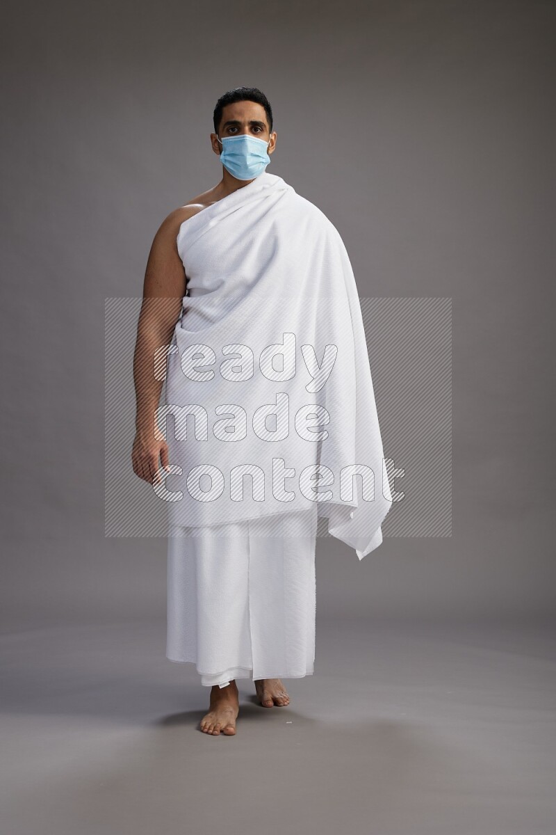 A man wearing Ehram with face mask Standing Interacting with the camera on gray background