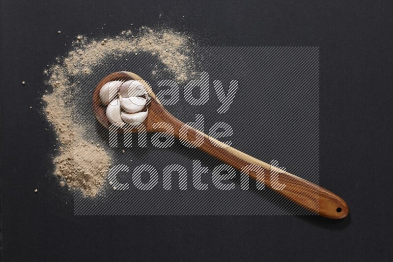 A wooden ladle full of garlic cloves with sprinkled powder on a black flooring