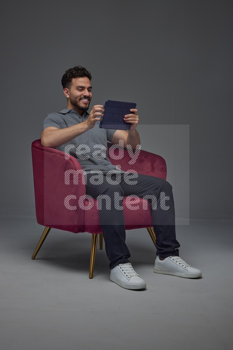 A man wearing casual and using his tablet while sitting on a burgundy chair eye level on a gray background