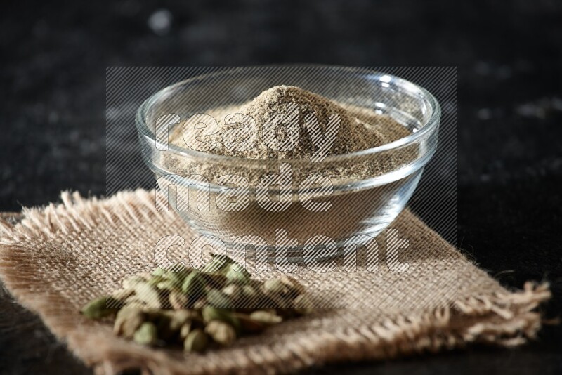 A glass bowl full of cardamom powder with cardamom seeds on a burlap piece on textured black flooring