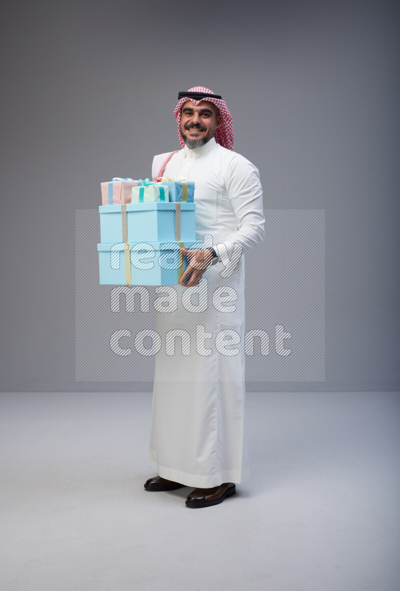 Saudi man Wearing Thob and red Shomag standing holding gift box on Gray background