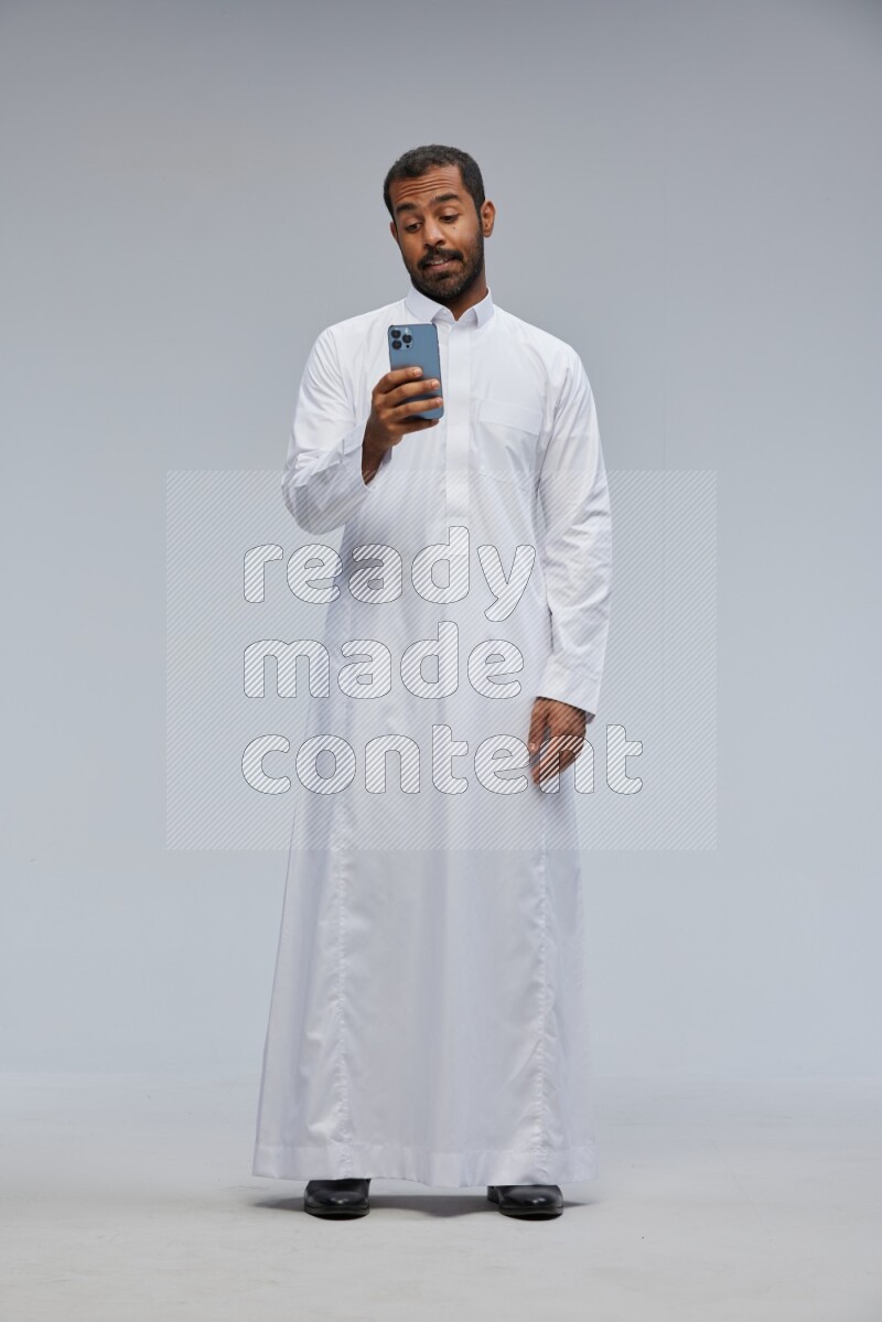 Saudi man wearing Thob standing texting on phone on Gray background
