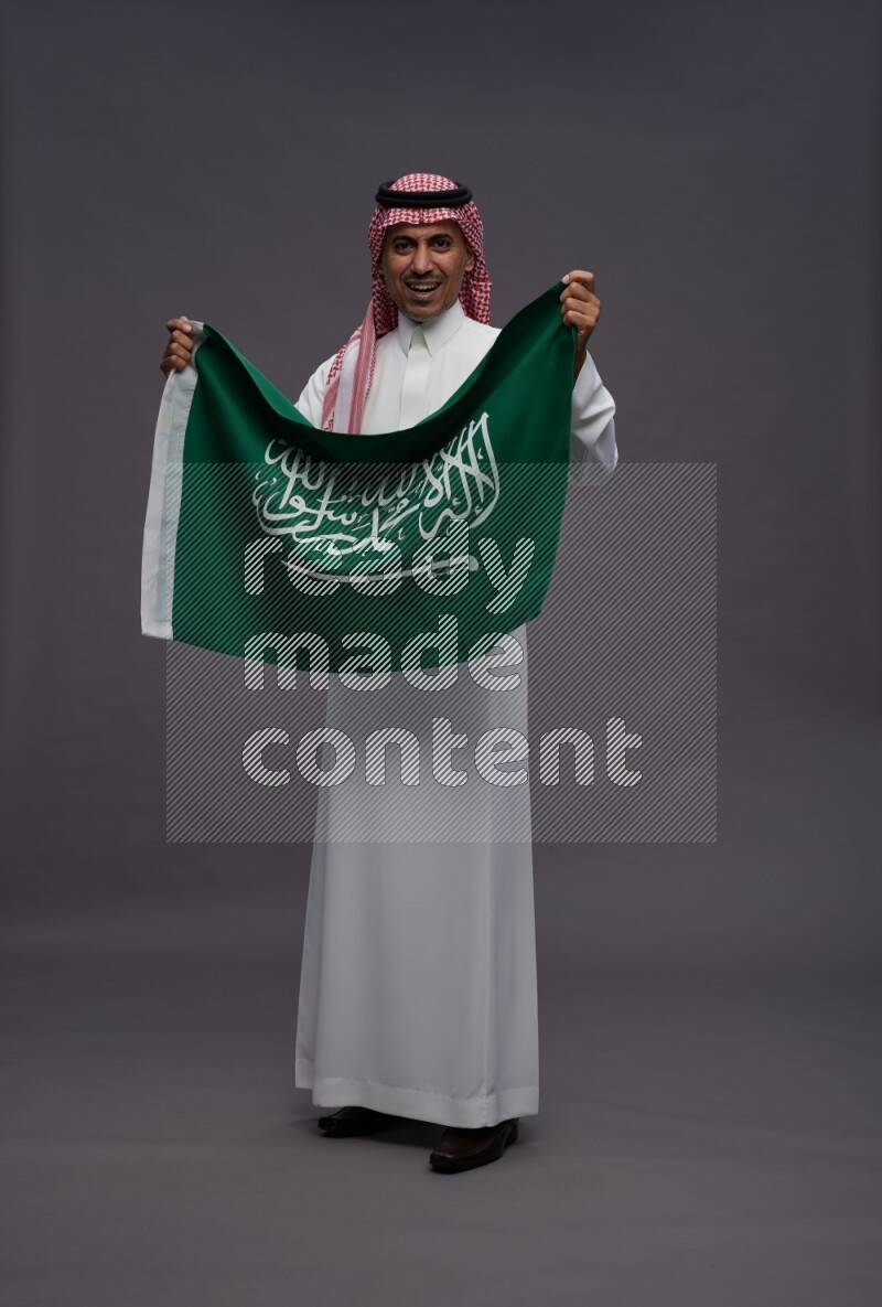 Saudi man wearing thob and shomag standing holding Saudi flag on gray background
