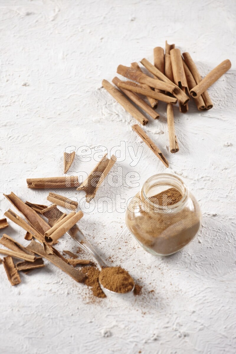 Herbal glass jar full cinnamon powder and a metal spoon surrounded by cinnamon sticks on a white background