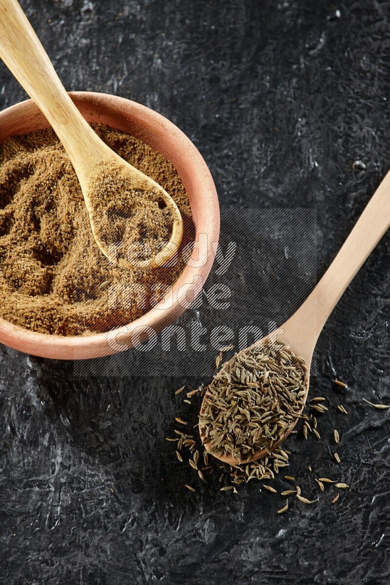 A wooden bowl and 2 wooden spoons full of cumin powder and cumin seeds on a textured black flooring