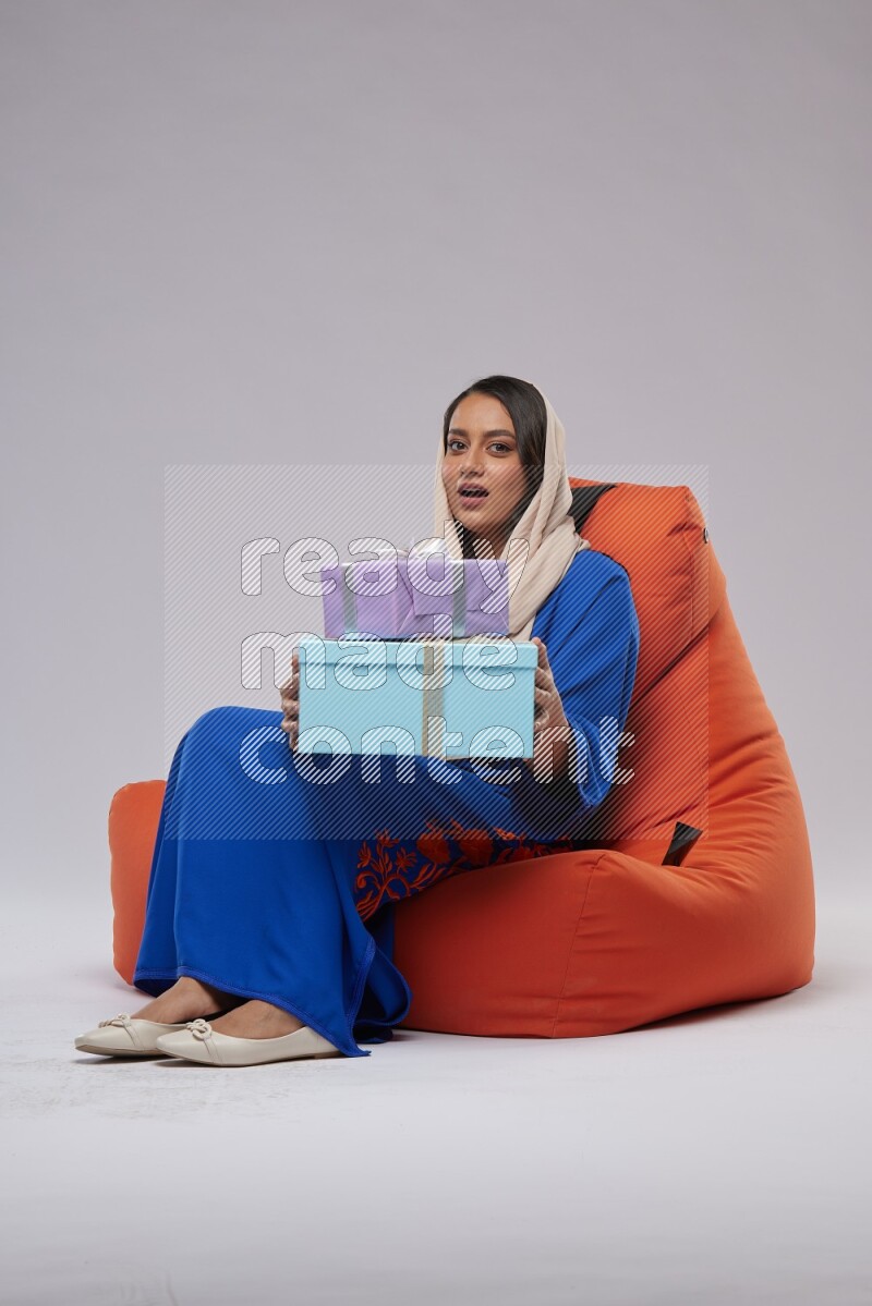A woman sitting on an orange beanbag wearing Jalabeya holding a gift box