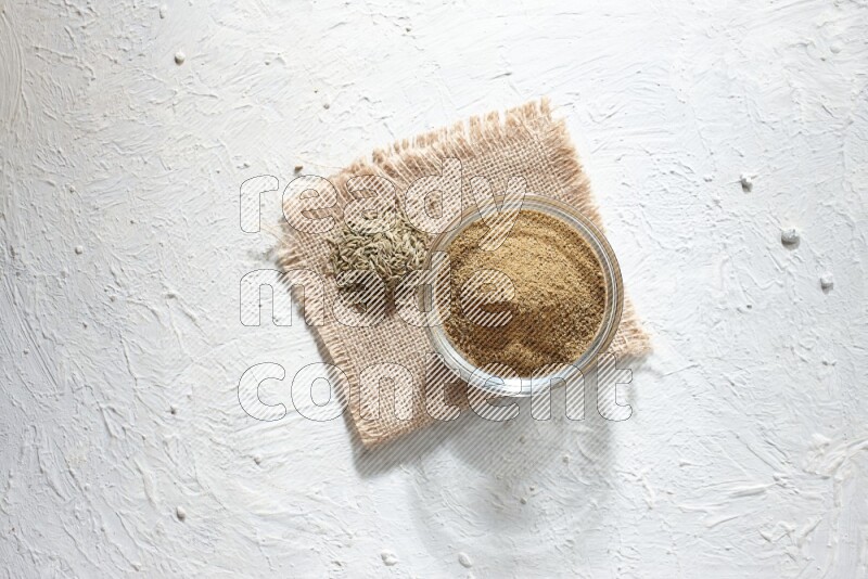 A glass bowl full of cumin powder with some of cumin seeds on burlap piece on a textured white flooring