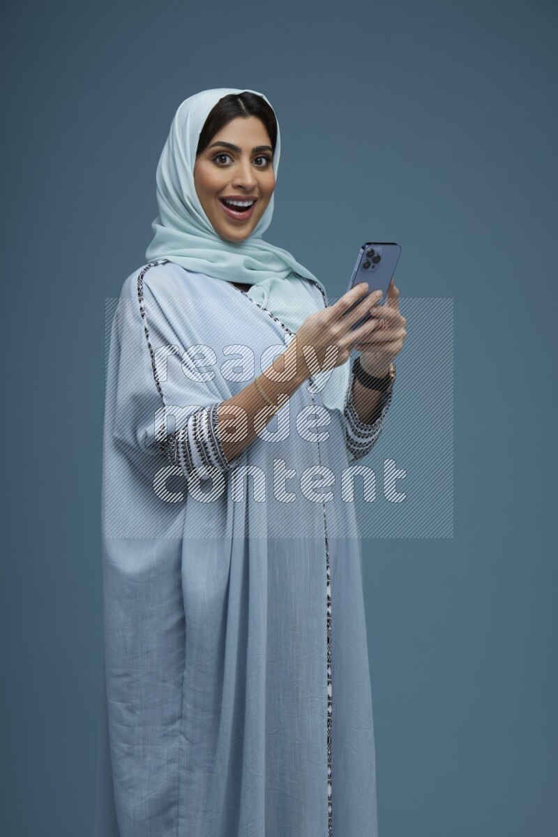 A Saudi woman posing with her phone in a blue background wearing a blue Abaya with hijab