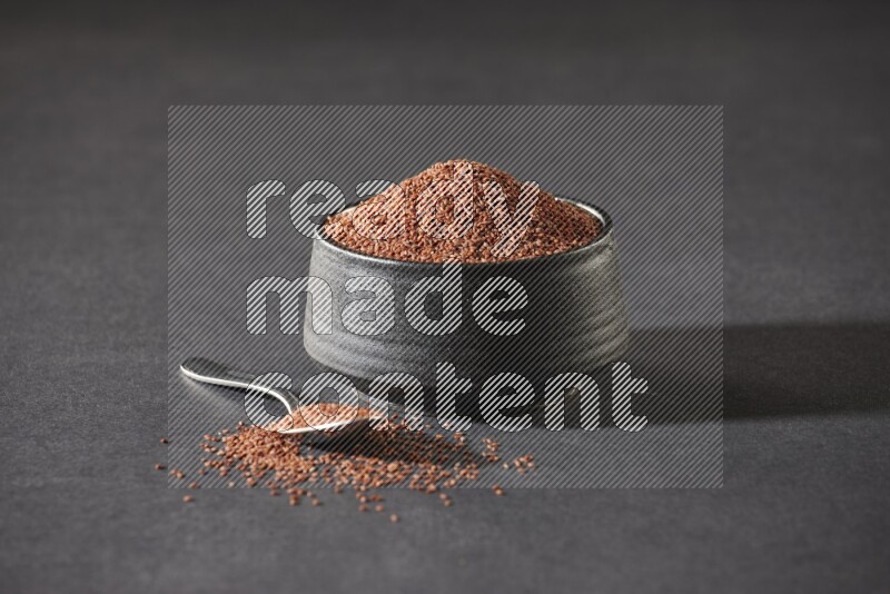 A black pottery bowl full of garden cress seeds and metal spoon full of the seeds on a black flooring