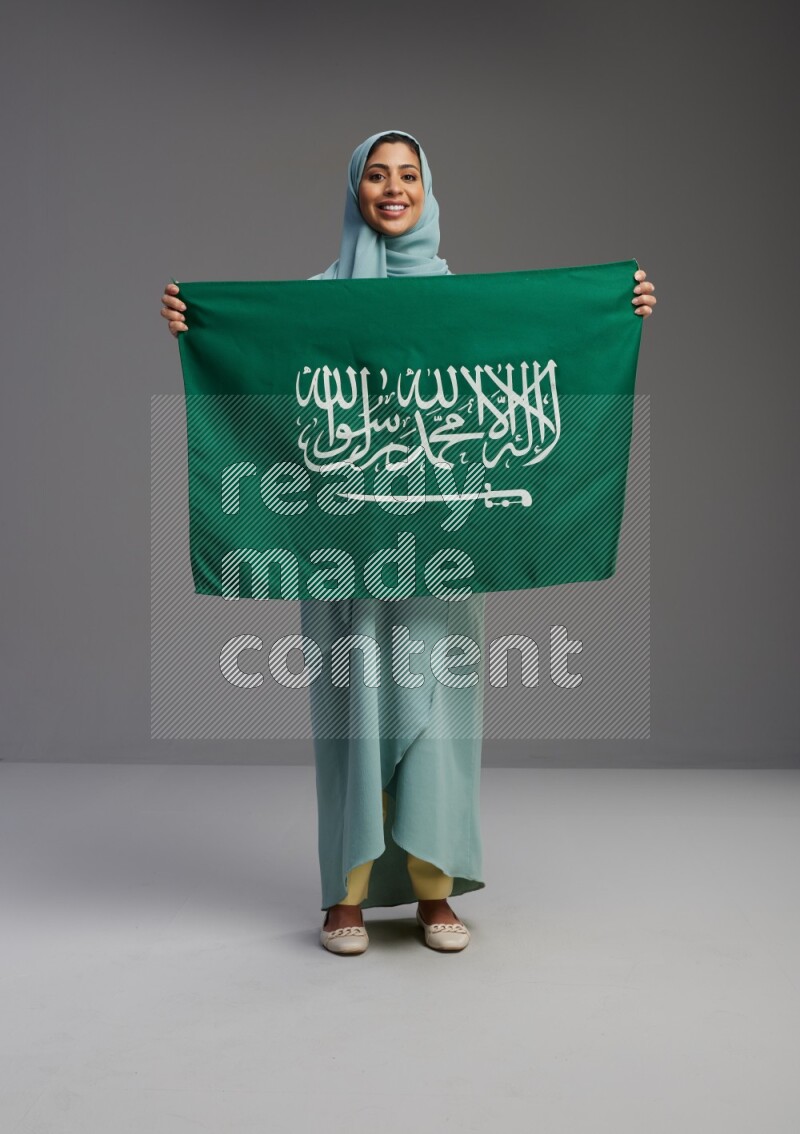 Saudi Woman wearing Abaya standing holding Saudi flag on Gray background