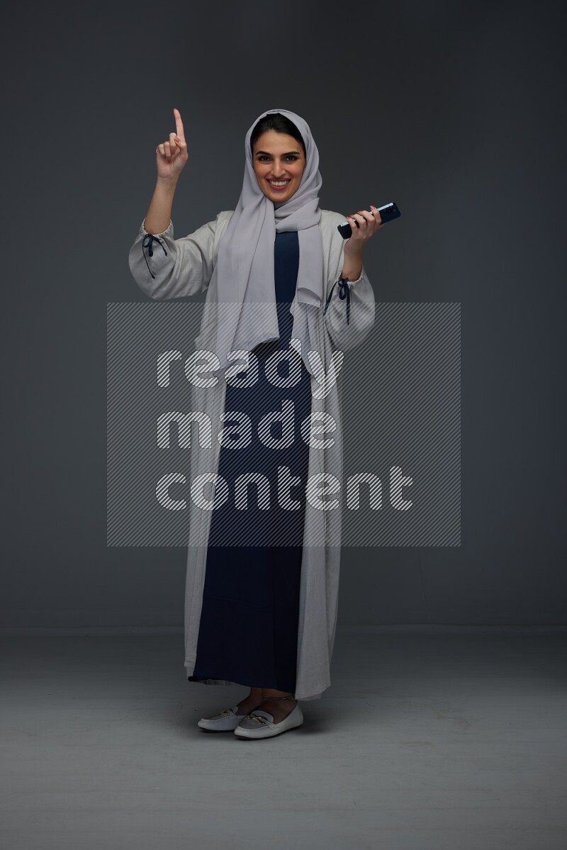 A Saudi woman wearing a light gray Abaya and head scarf standing and holding a phone on a grey background