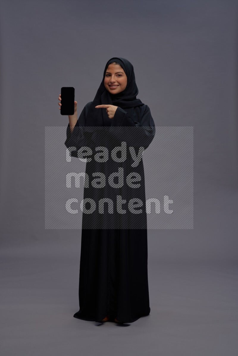 Saudi woman wearing Abaya standing showing phone to camera on gray background