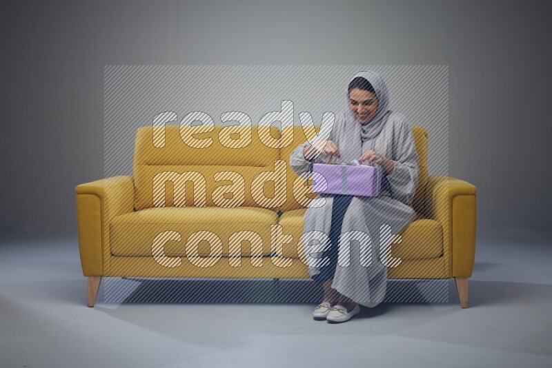 A Saudi woman wearing a light gray Abaya and head scarf sitting on a yellow sofa holding a gift box beside multi color gift boxes eye level on a grey background