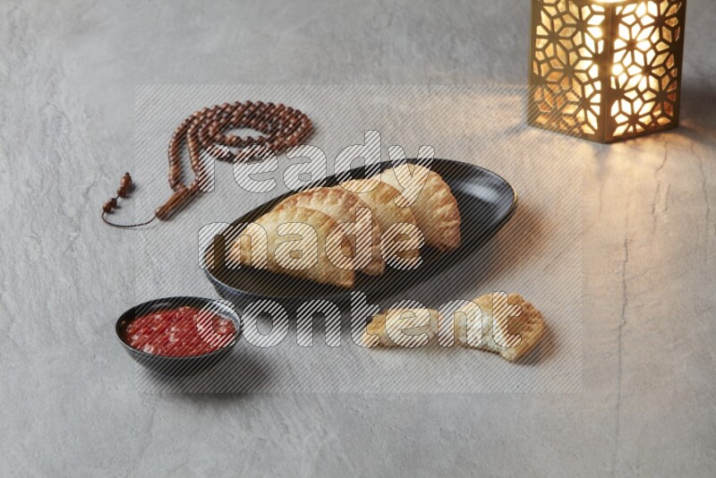 Four fried sambosas in an oval shaped black plate, beside a cut cheese sambosa, a brown misbaha and a golden lantern on a gray background