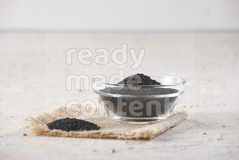 A glass bowl full of black seeds on a burlap piece on textured white flooring