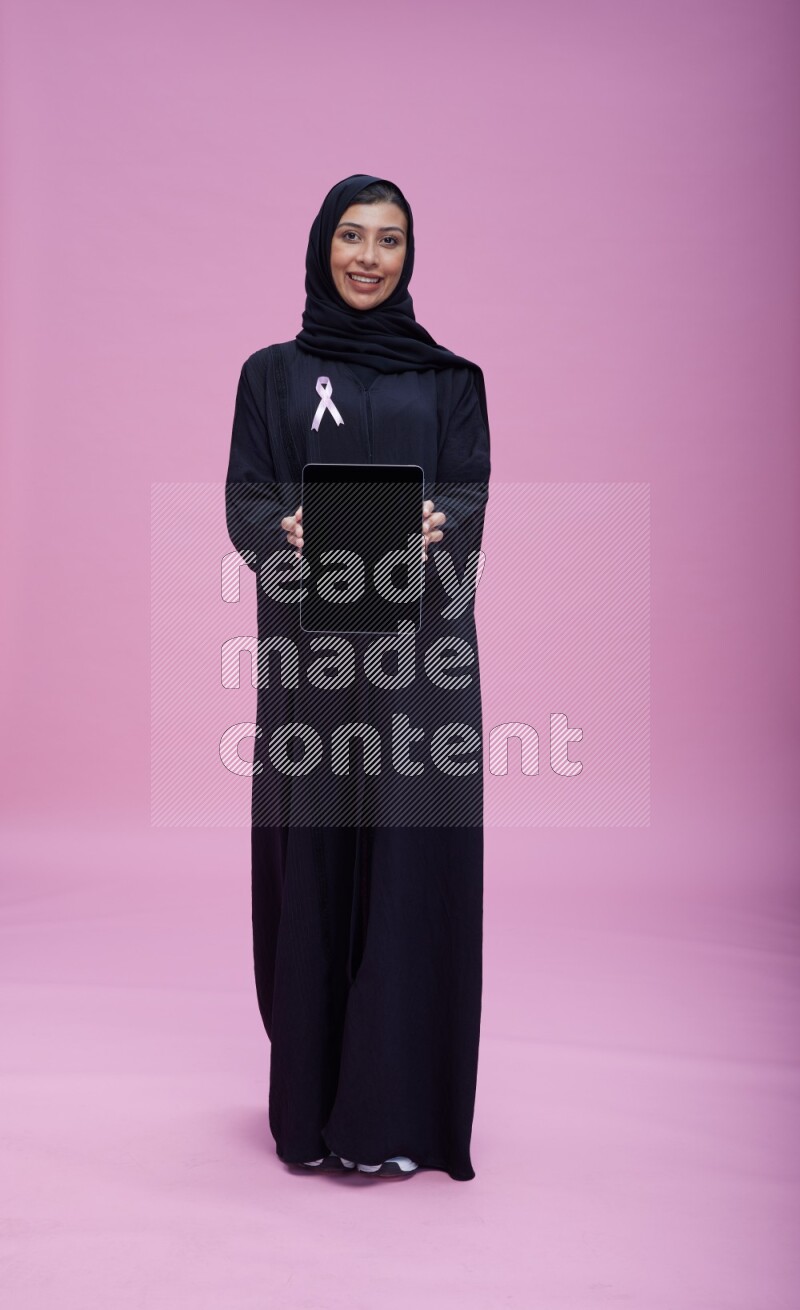 Saudi woman wearing pink ribbon on Abaya standing showing a tablet to camera on pink background