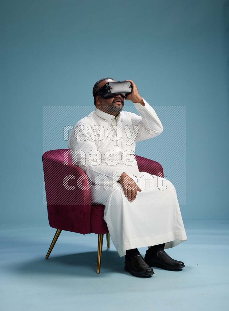 Saudi Man without shimag sitting on chair  gaming with VR on blue background