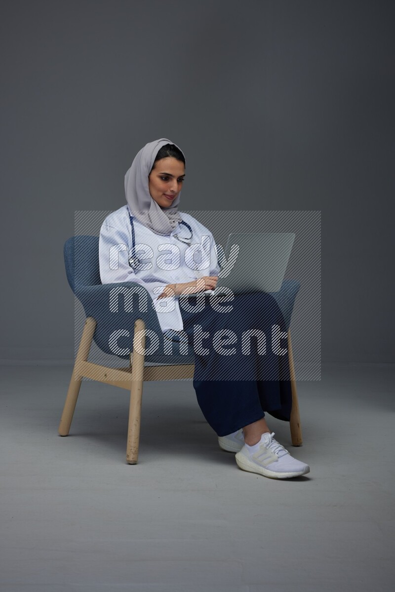 A doctor wearing a light gray head scarf sitting on a dark grey chair and using a laptop eye level on a grey background