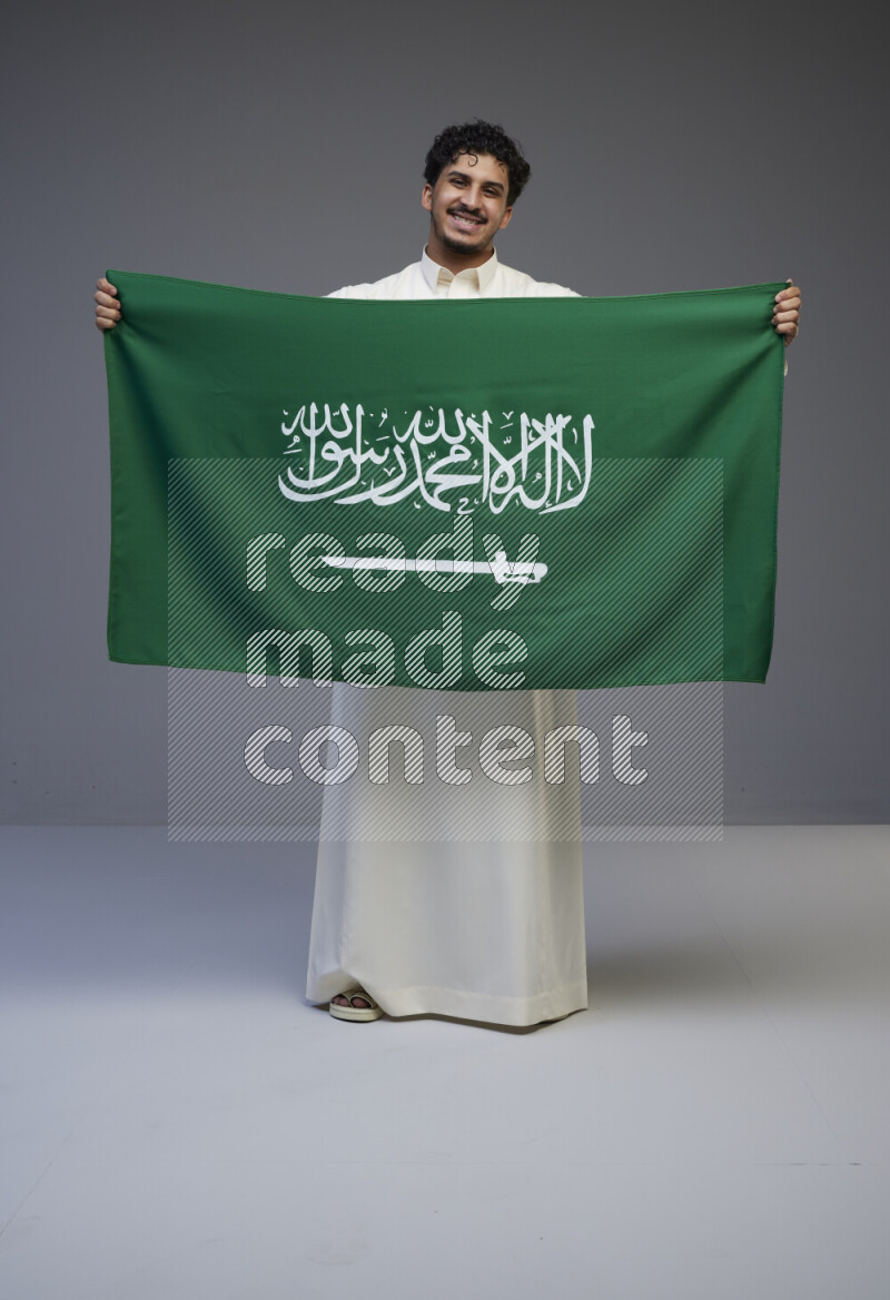 A Saudi man standing wearing thob holding big Saudi flag on gray background