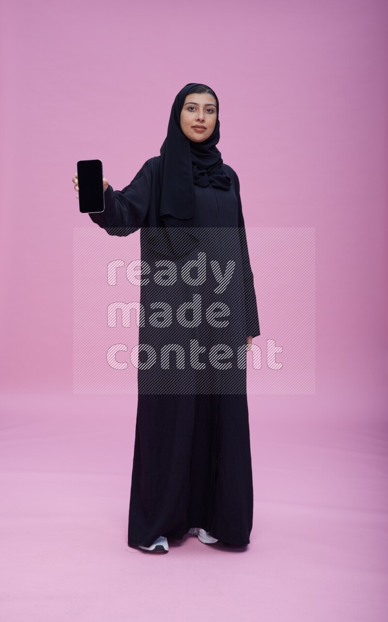 Saudi woman wearing Abaya standing showing phone to camera on pink background