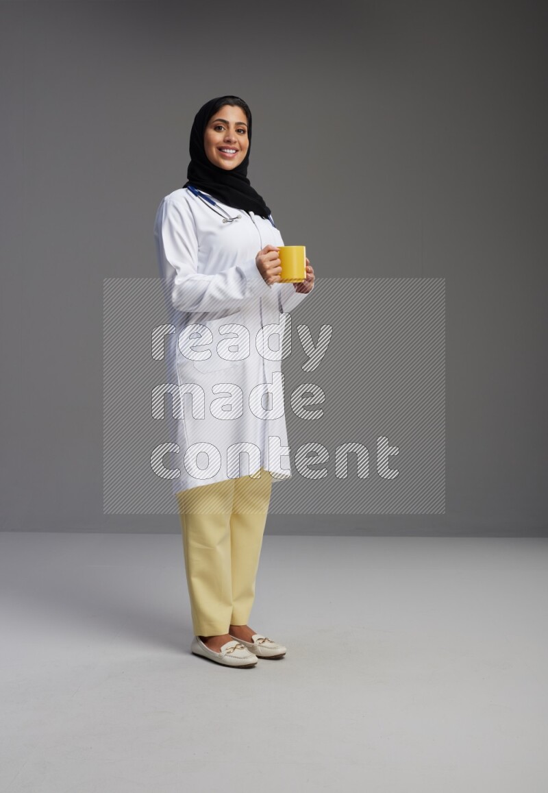Saudi woman wearing lab coat with stethoscope standing holding mug on Gray background