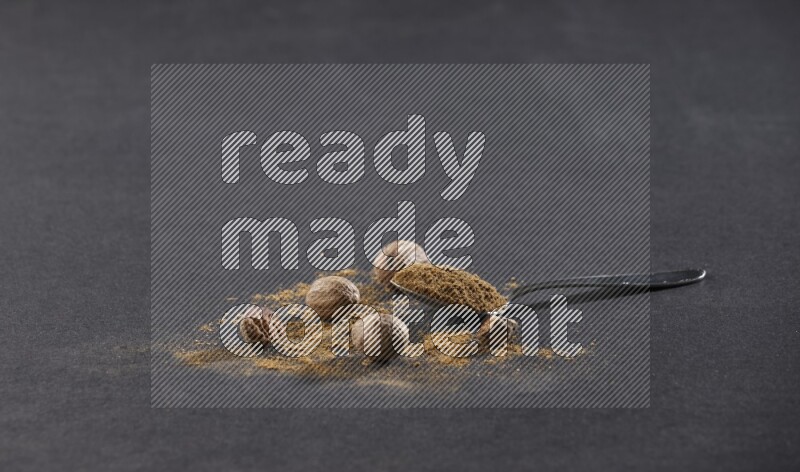 A metal spoon full of nutmeg powder with nutmeg powder and whole seeds beside it on a black flooring