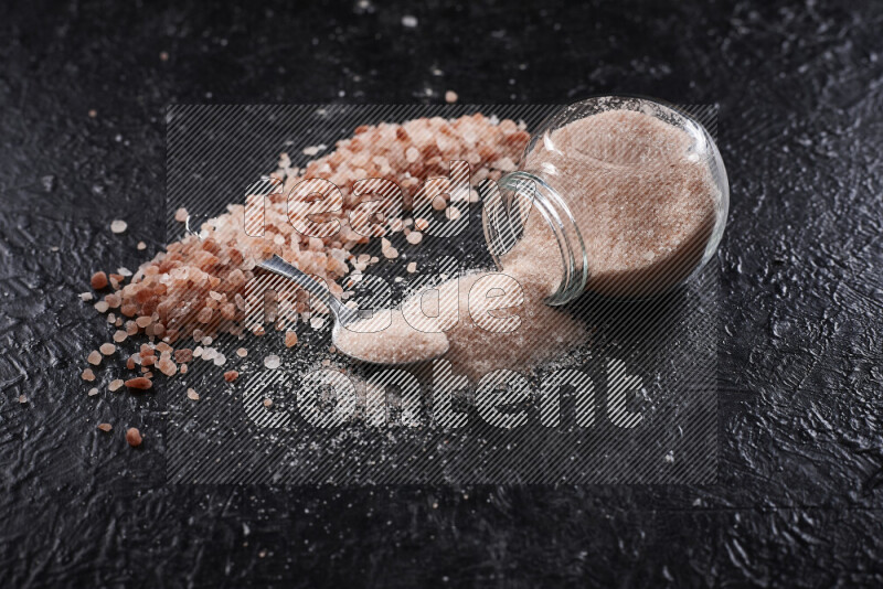 A glass jar full of fine himalayan salt with some himalayan crystals beside it on a black background