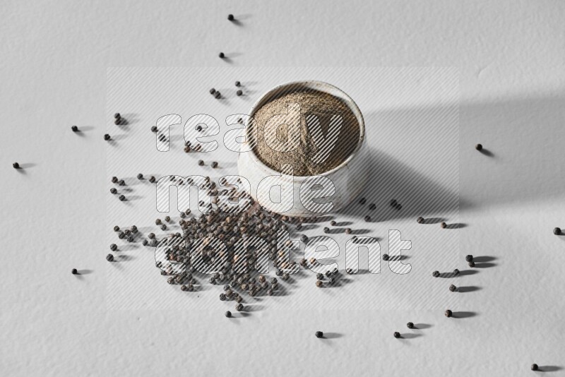 A white ceramic bowl full of black pepper powder and black pepper beads spread on white flooring