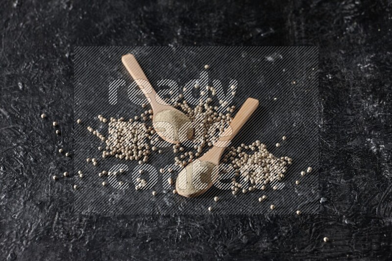 2 wooden spoons full of white pepper powder with white pepper beads on textured black flooring