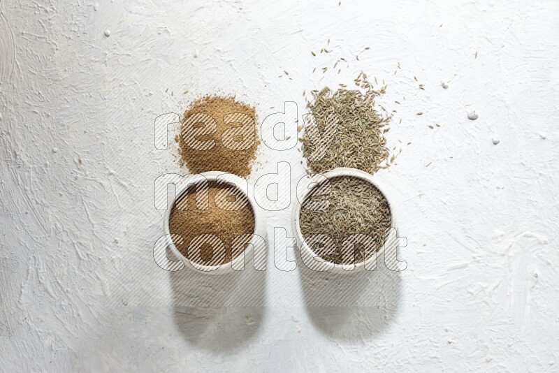 2 beige bowls full of cumin seeds and powder with spilled powder and seeds on textured white flooring