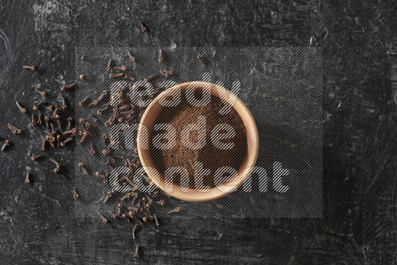 A wooden bowl full of cloves powder on a textured black flooring