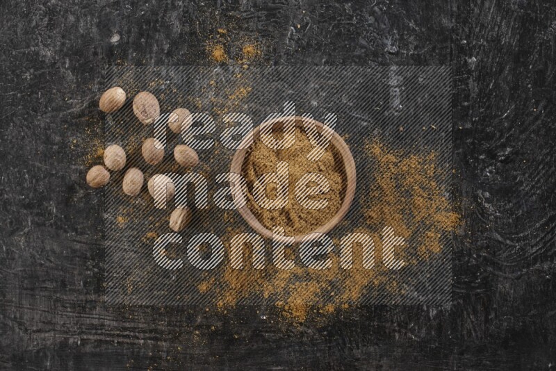 A wooden bowl full of nutmeg powder with whole seeds and sprinkled powder beside it on a textured black flooring