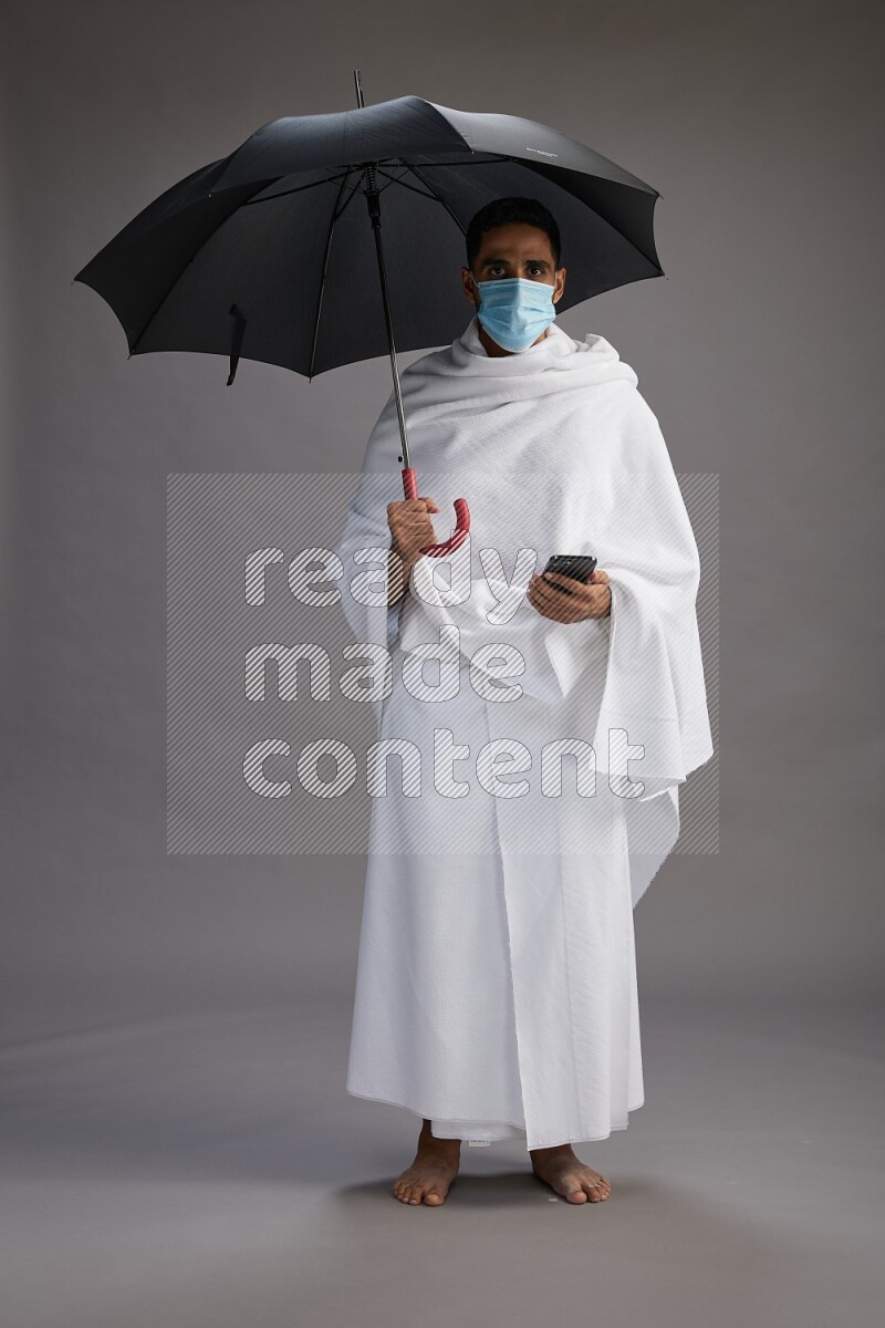 A man wearing Ehram and mask Standing holding umbrella on gray background on gray background