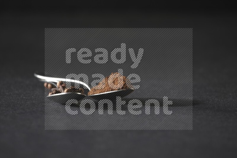 2 metal spoons, one full of cloves grains and the other full of cloves powder on a black flooring