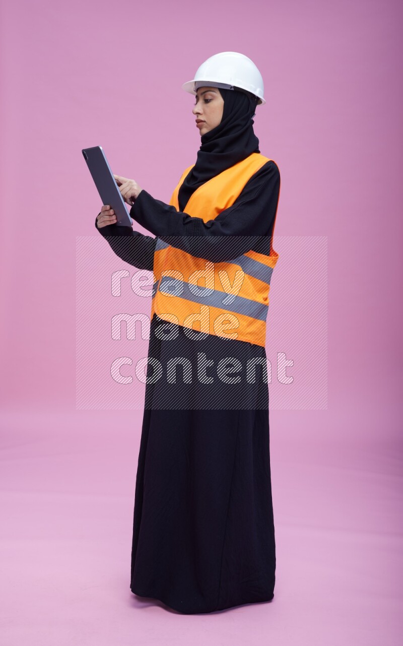 Saudi woman wearing Abaya with engineer vest and helmet standing working on tablet on pink background