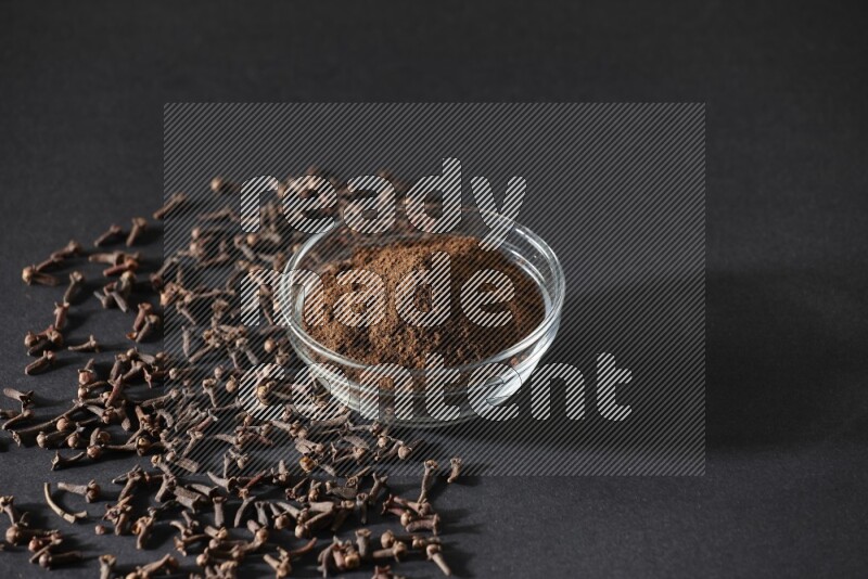 A glass bowl full of cloves powder with cloves grains spread on black flooring