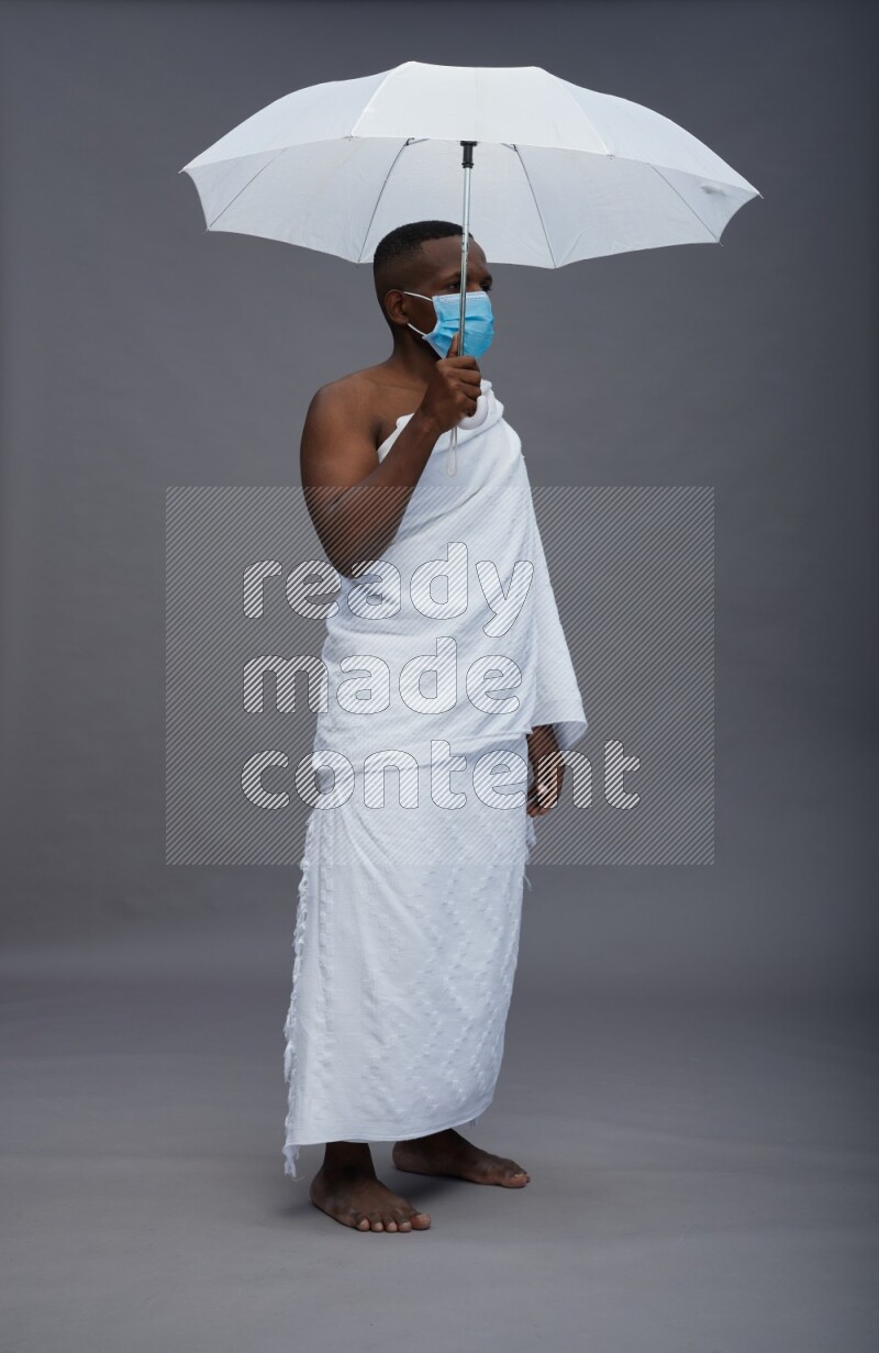 A man wearing Ehram with face mask Standing holding umbrella on gray background