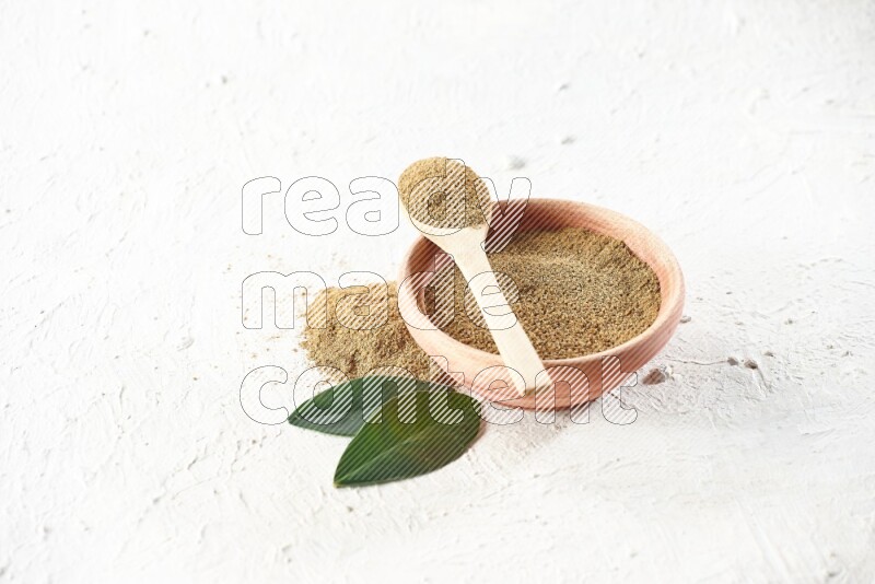 A wooden bowl and wooden spoon full of cumin powder on textured white flooring