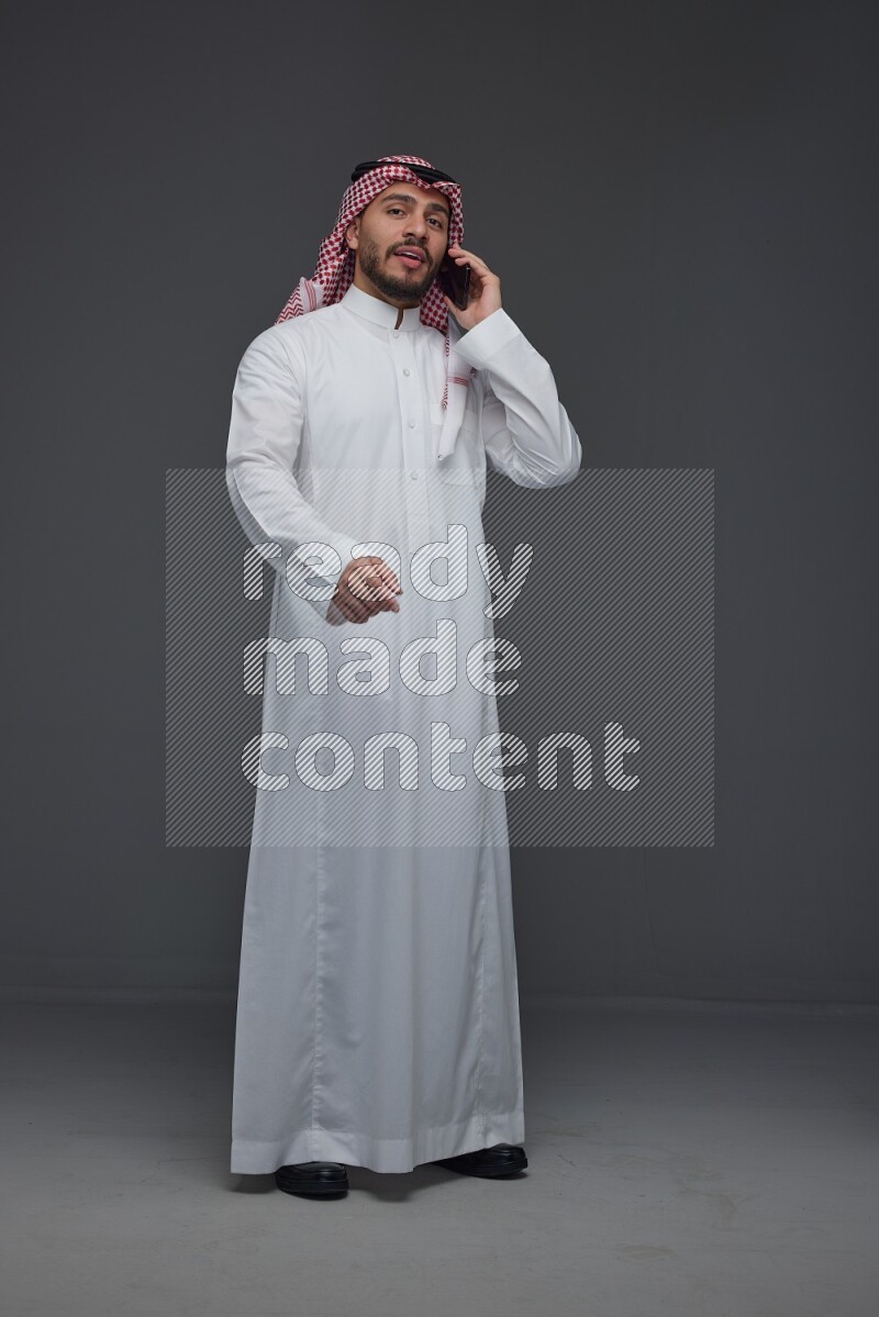 A Saudi man wearing Thobe and Shmagh talking in the phone eye level on a gray background