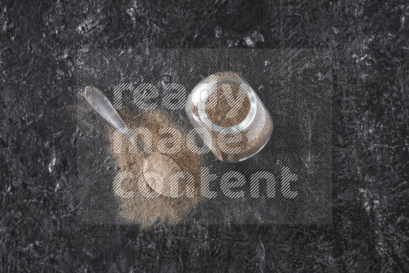 A glass spice jar full of black pepper powder and a metal spoon full of powder on textured black flooring