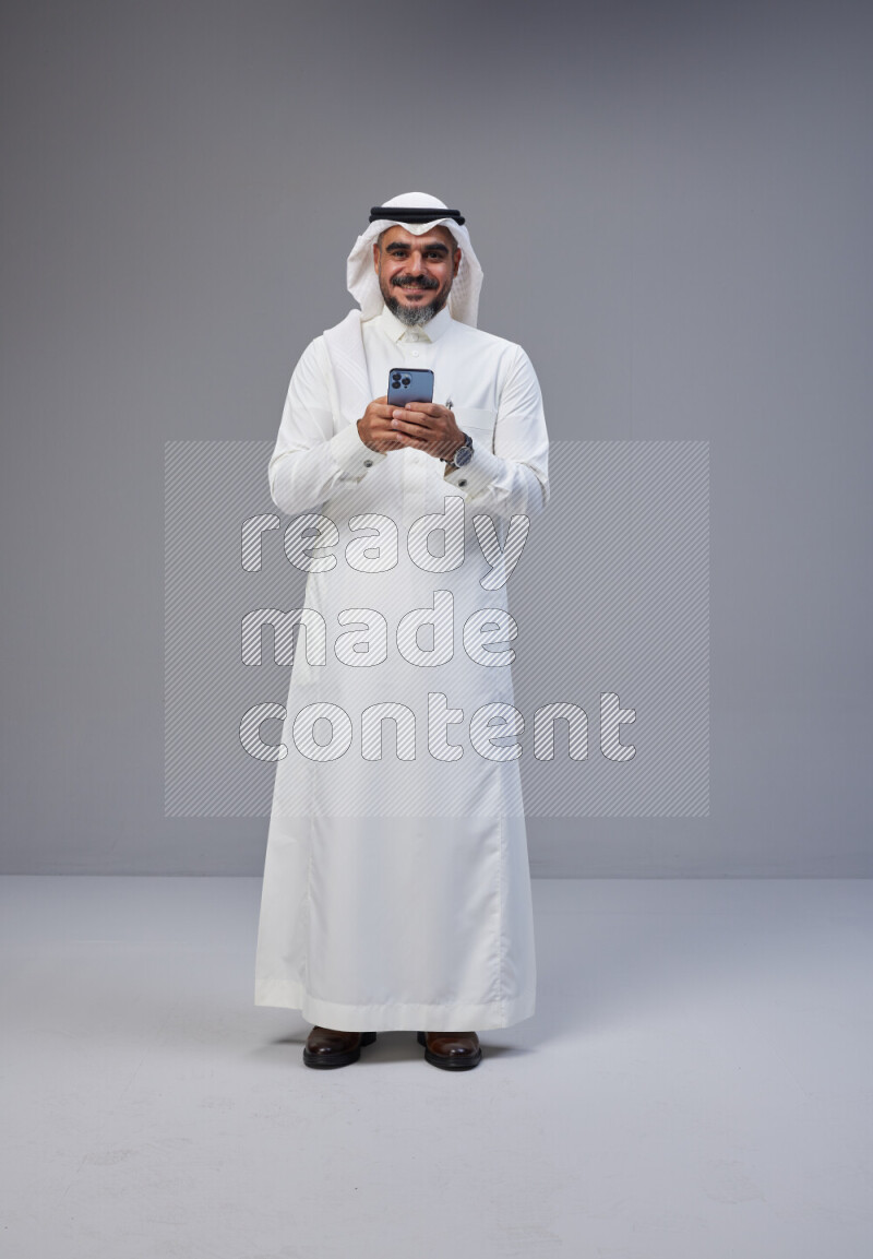 Saudi man Wearing Thob and white Shomag standing texting on phone on Gray background