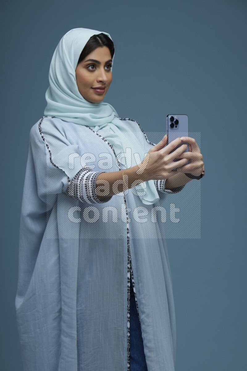 A Saudi woman Taking a Selfie on a blue background wearing a blue Abaya with hijab