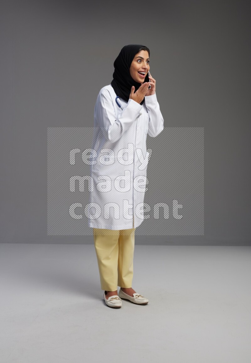 Saudi woman wearing lab coat with stethoscope standing talking on phone on Gray background