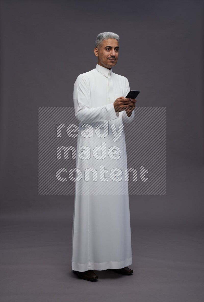 Saudi man wearing thob standing texting on phone on gray background
