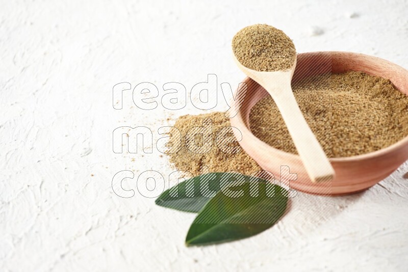 A wooden bowl and wooden spoon full of cumin powder on textured white flooring