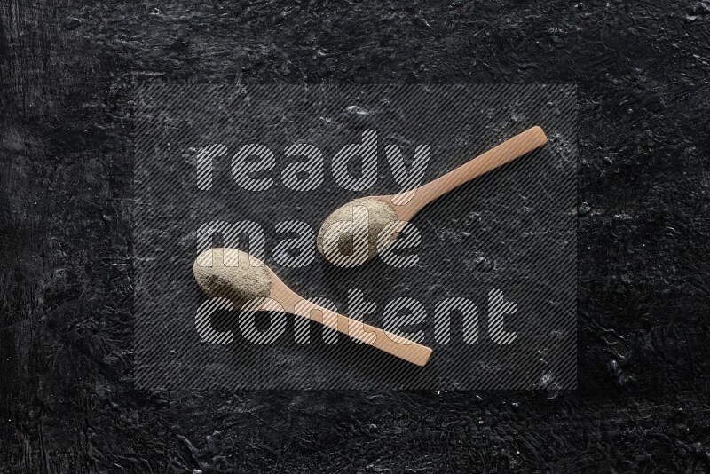 2 wooden spoons full of white pepper powder on textured black flooring