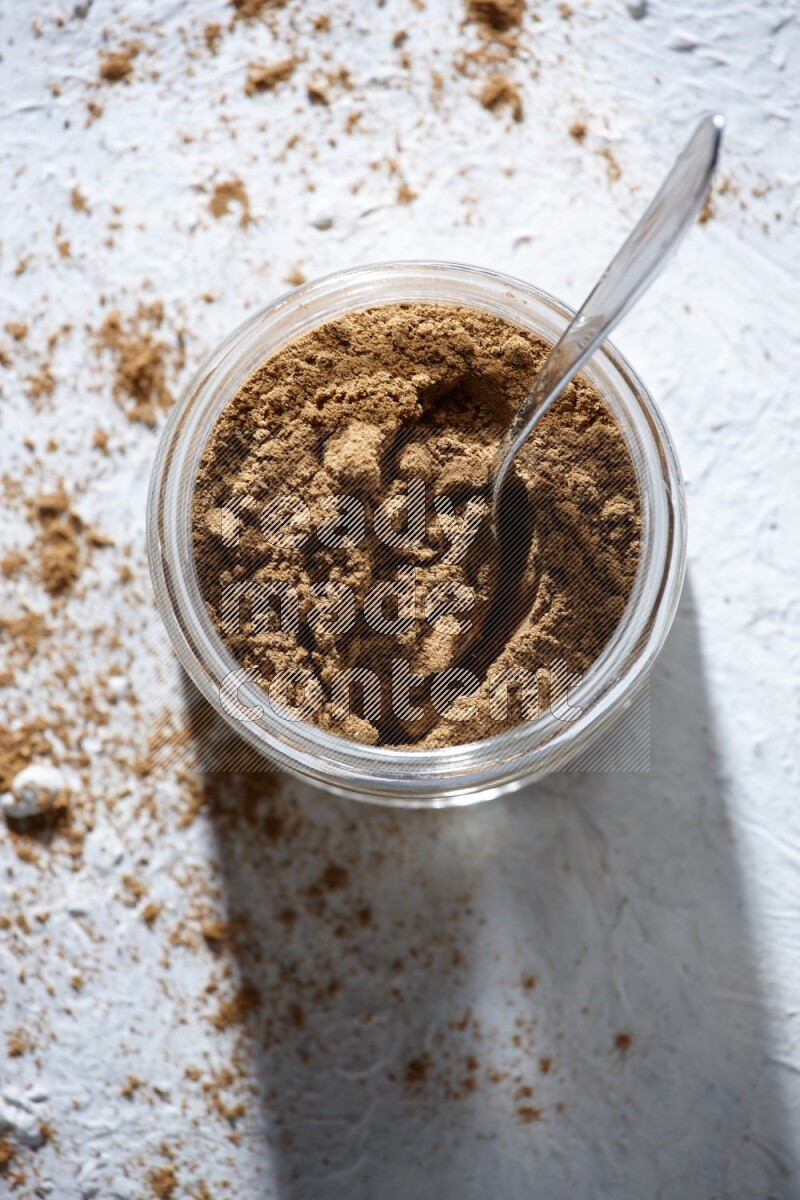 A glass jar and a metal spoon full of allspice powder on a textured white flooring
